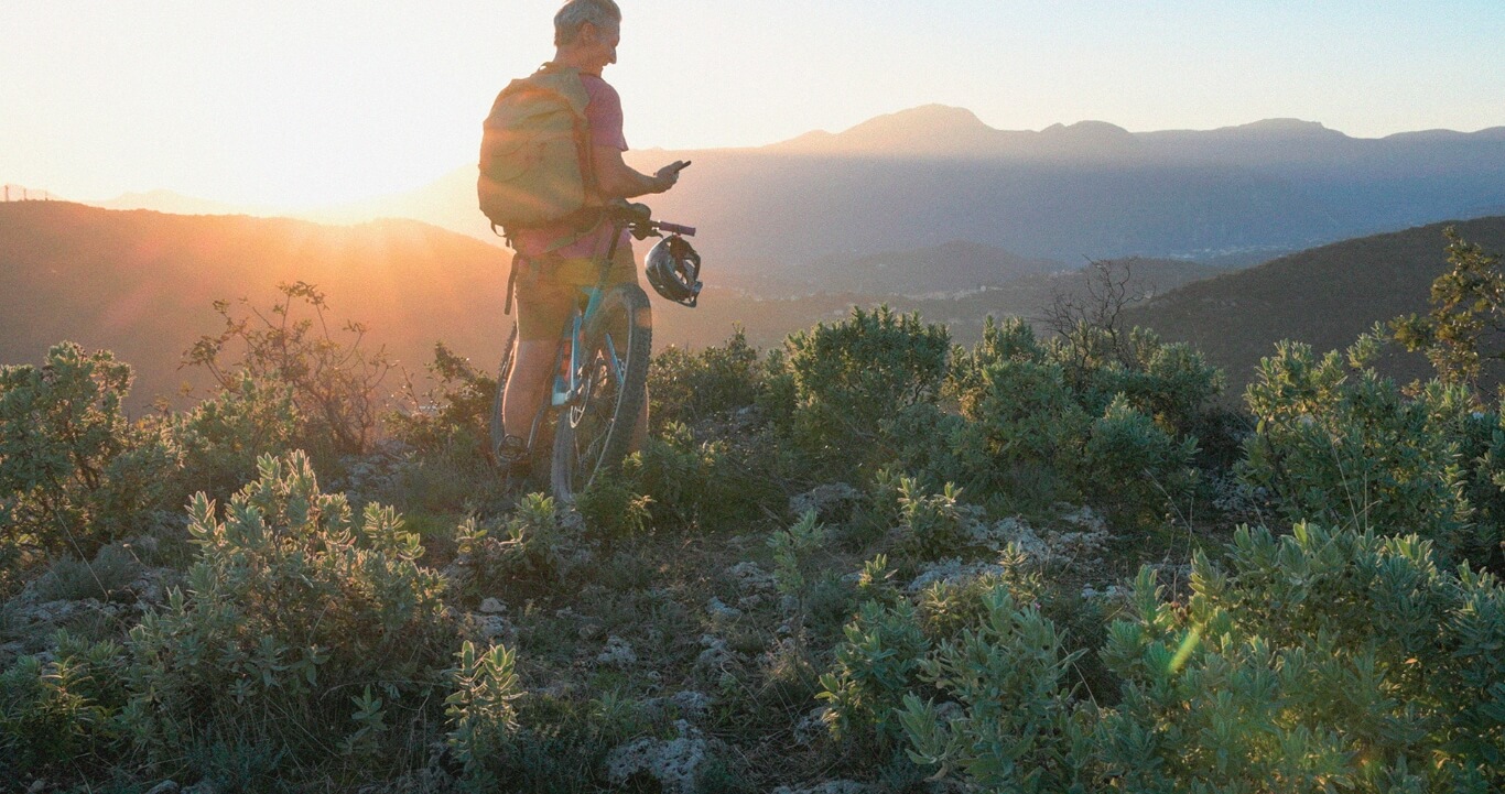 man stops on a cliff ledge to overlook mountain scape during sunrise and look at his phone