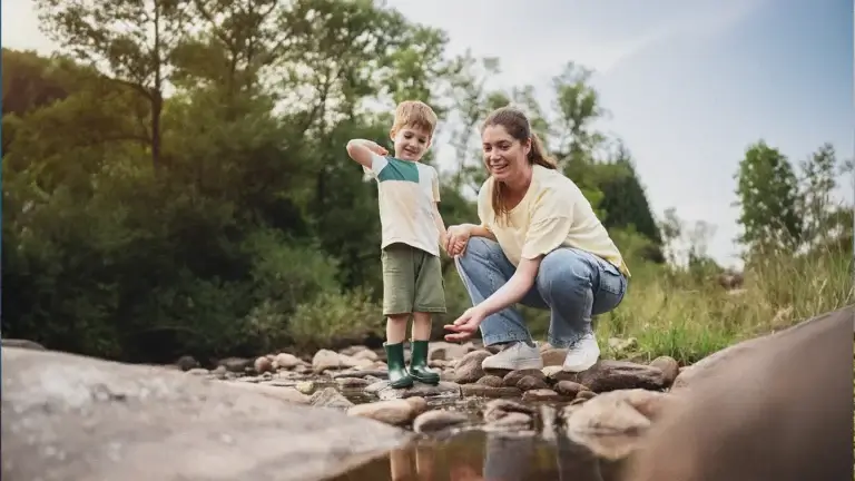 A woman and young child explore a shallow rocky creek in a natural outdoor setting in their family adventures in Big Sky, Montana. The child stands in green rain boots while the woman crouches beside them, both focused on the water below. Surrounded by trees, grasses, and smooth river stones, the scene captures a quiet moment of connection and outdoor adventure in a peaceful landscape.