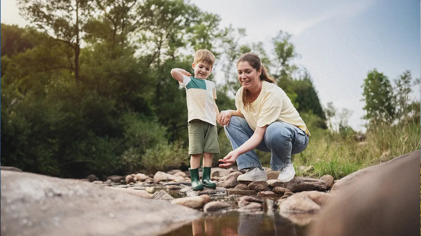A woman and young child explore a shallow rocky creek in a natural outdoor setting in their family adventures in Big Sky, Montana. The child stands in green rain boots while the woman crouches beside them, both focused on the water below. Surrounded by trees, grasses, and smooth river stones, the scene captures a quiet moment of connection and outdoor adventure in a peaceful landscape.