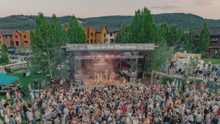 Crowd enjoying a live outdoor concert during the Music in the Mountains summer festival in Big Sky, Montana. The stage is surrounded ny trees and the mountains can be seen in the distance.