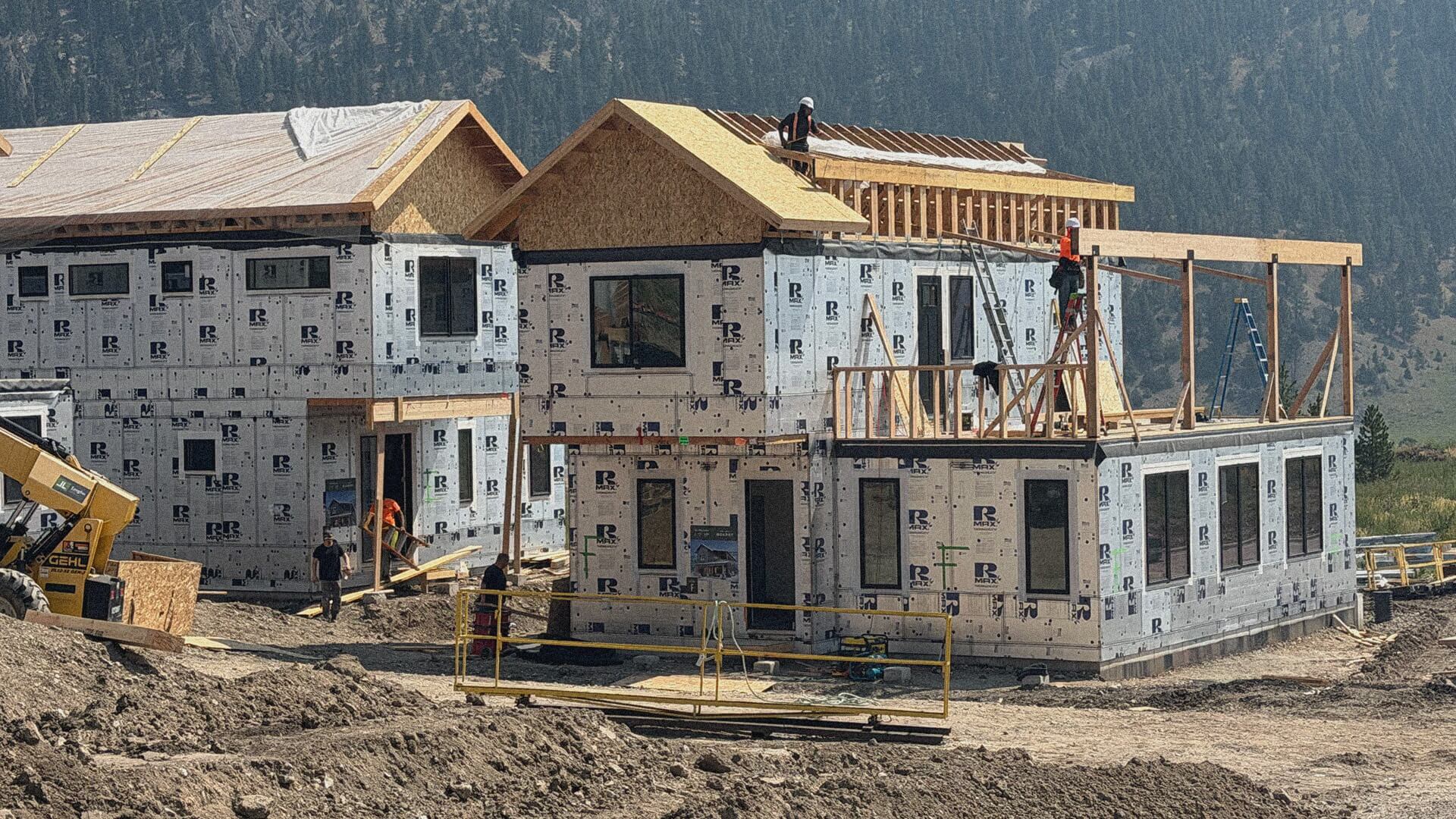 New homes in Big Sky are under construction at Big Sky Quarry, with workers framing the roofs against a dramatic Montana mountain backdrop.